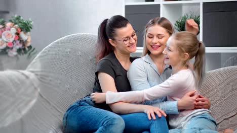 Same-sex-female-family-smiling-hugging-daughter-sitting-on-couch-looking-at-camera