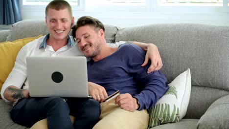 Gay-couple-relaxing-on-couch-using-laptop-computer.-Talking-and-holding-card.