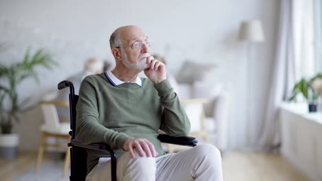 Tracking-medium-shot-of-disabled-senior-man-in-eyeglasses-sitting-in-wheelchair-looking-away-and-thinking-in-assisted-living-home