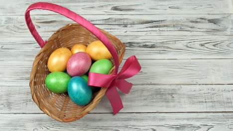 Male-hand-puts-a-gift-basket-with-colorful-chicken-eggs-on-a-wooden-white-background,-Easter-holiday,-copy-space,-celebration