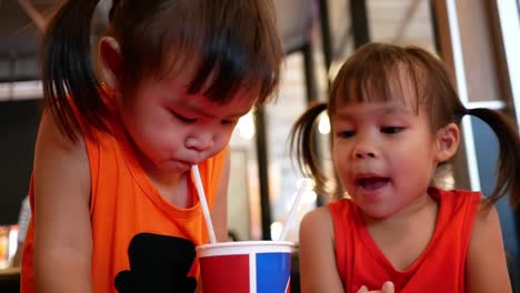 Little-Asian-child-girl-with-her-sister-sipping-her-drink-together-while-sitting-wait-food-in-restaurant.-Selective-focus.