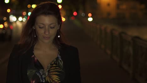 Bonita-feliz-mujer-moderna-caucásica-usando-vestido-de-flores,-chaqueta-negra-y-pelo-rojo-caminando-por-la-calle-y-escribiendo-un-mensaje-de-texto-en-su-teléfono-inteligente-por-la-noche.-Paris-4K-UHD.-A-cámara-lenta.