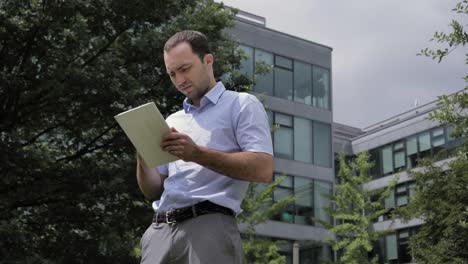 Geschäftsmann-steht-im-Stadtpark,-in-der-Nähe-von-Bürogebäuden-mit-Tablet