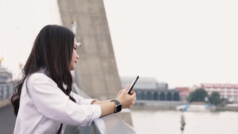 Porträt-von-attraktiven-jungen-asiatischen-Frau-mit-Smartphone-Chat-mit-Freunden-und-Surfen-in-sozialen-Medien-auf-einem-Handy,-während-auf-der-Brücke-stehen.