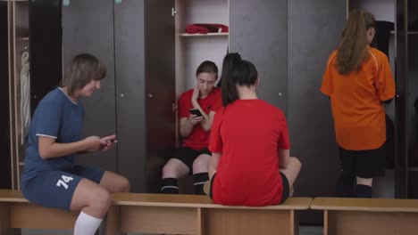 Jugadoras-de-fútbol-femeninas-usando-teléfonos-inteligentes-y-hablando-en-la-sala-de-lockers