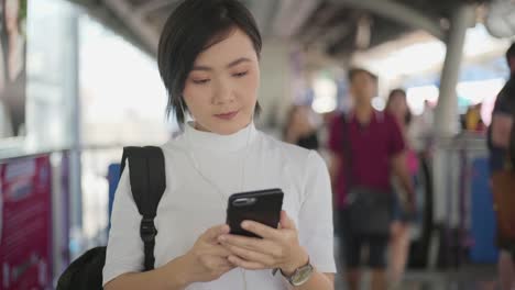 Retrato-de-mujer-asiática-con-auriculares-escuchando-música-y-usando-teléfono-inteligente-para-chatear-con-amigos-o-navegar-mientras-espera-un-tren.-Tecnología-en-la-vida-cotidiana-y-los-viajes.