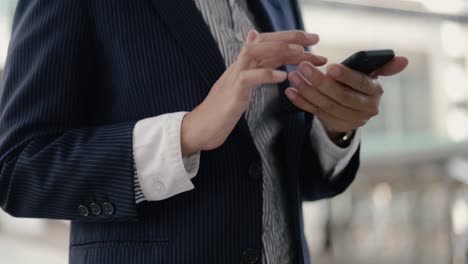 Cerrar-a-la-mujer-de-negocios-asiática-en-traje-negro-está-utilizando-un-teléfono-inteligente-que-envía-mensajes-compartiendo-mensajes-en-las-redes-sociales-mientras-está-de-pie-en-la-calle-cerca-de-un-gran-edificio-de-oficinas-urbana-con-puesta-de-so