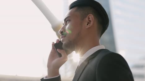 Asian-businessman-in-a-black-suit-using-his-smartphone-talking-on-the-phone-while-standing-outside-on-the-street-near-a-big-office-building-urban-having-sunlight-sunset.