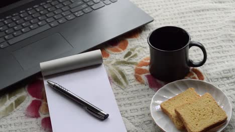 Breakfast-coffee-in-morning-sunlight-with-laptop-computer-black-color-pen-and-white-ruled-paper-notebook,-ceramic-cup-saucer-and-biscuit-on-top-office-place-working-desk-background.-Lifestyle-image.