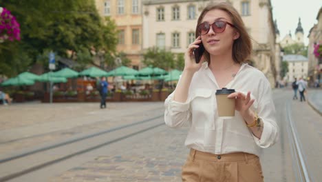 Das-Mädchen-telefoniert.-Sie-geht-und-lächelt.-Sie-hält-eine-Tasse-Kaffee.-Sie-trägt-eine-Sonnenbrille.-Die-Menschen-gehen-im-Hintergrund.-4K