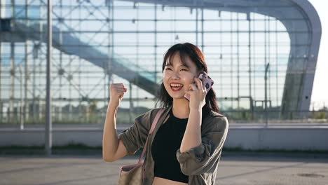 Appealing-cheerful-asian-young-woman-talking-on-smartphone-on-the-background-of-airport-building