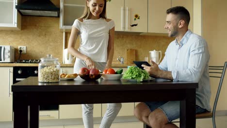 Inclinación-hacia-arriba-de-la-atractiva-pareja-charlando-en-la-cocina-temprano-en-la-mañana.-Hombre-guapo-con-tablet-mientras-su-novia-cocina