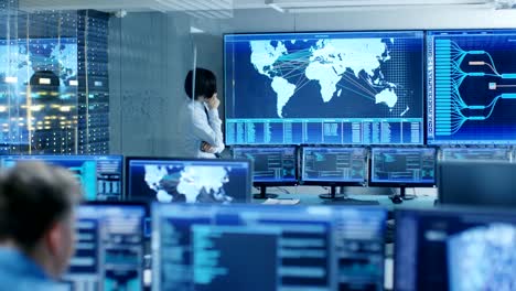 In-the-System-Control-Room-Chief-Engineer-Thinks-While-Standing-Before-Big-Screen-with-Interactive-Map-on-it.-Data-Center-is-Full-of-Monitors-Showing-Graphics.