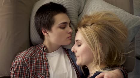 Two-young-lesbian-girls-are-lying-on-the-bed,-a-girl-with-short-hair-kisses-her-partner-in-the-forehead,-an-LGBT-family-concept,-they-sleep.-60-fps