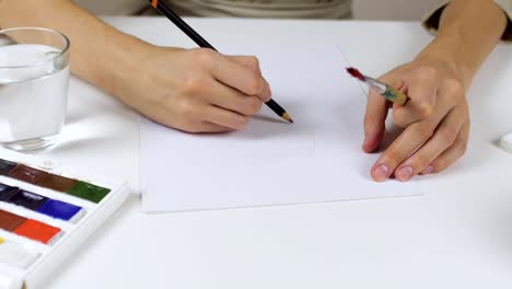 Unrecognizable-woman-draws-a-pencil-sketch-on-paper-Easter-card.-On-the-table-is-a-palette-with-paints-and-a-glass-of-water.