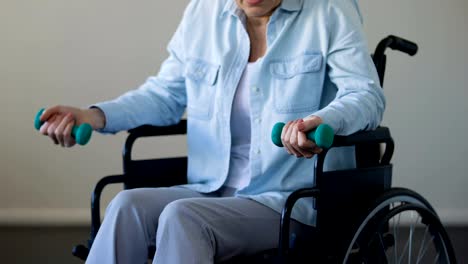 Mature-lady-sitting-in-wheelchair-and-trying-to-lift-dumbbells,-rehabilitation