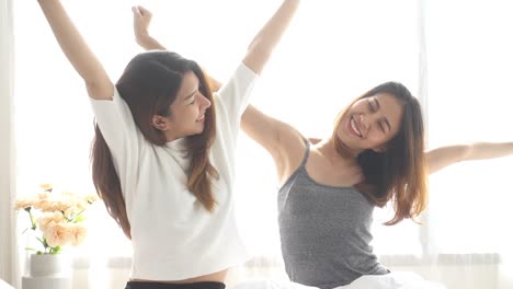 Asian-women-lesbian-happy-couple-waking-up-in-morning.-Asian-girls-sitting-on-bed-stretching-in-cozy-bedroom-at-home.-Funny-women-friends-after-wake-up.-LGBT-Lesbian-couple-together-indoors-concept.