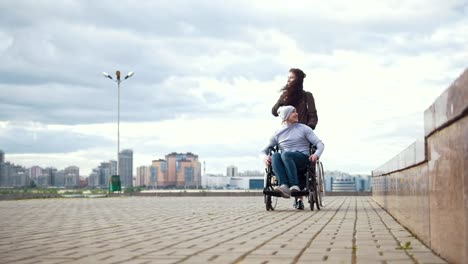 Disabled-man-in-a-wheelchair-with-young-woman-walking-at-the-city-street