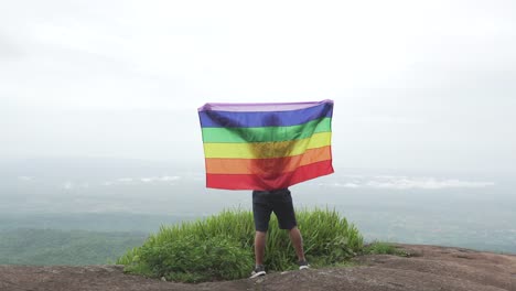 Mann-erhöhen-Regenbogen-Farbe-LGBTI-Fahnenschwingen-in-harten-Wind-auf-Mountain-Top-Sicht