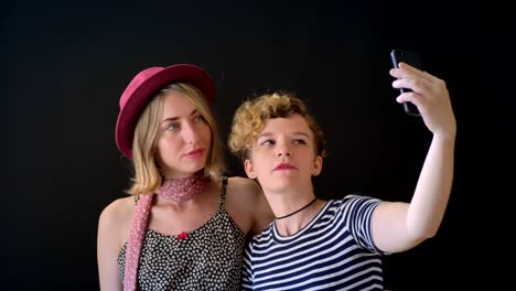 Two-pretty-young-blonde-women-embracing-and-taking-selfie,-isolated-on-black-studio-background