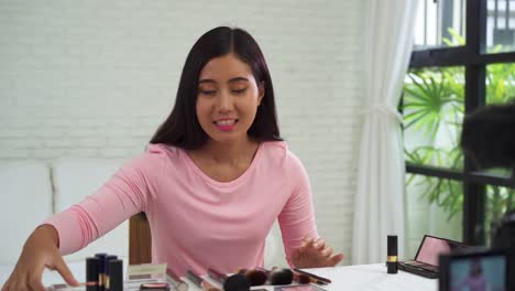 Blogger-de-belleza-actualmente-cosméticos-de-belleza-sentada-en-frente-la-cámara-para-la-grabación-de-vídeo.-Hermosa-mujer-use-cepillo-mientras-revisión-maquillaje-tutorial-transmiten-video-en-vivo-en-red-social-por-internet.