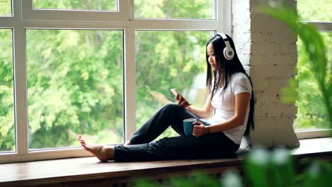 Pretty-Asian-student-is-enjoying-music-in-headphones-sitting-on-window-sill-and-using-smartphone-touching-screen.-Modern-lifestyle-and-technology-concept.