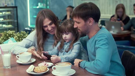 Family-in-a-cafe,-dad,mom,-daughter-at-the-table-watching-something-on-the-phone-and-discuss,-smile.