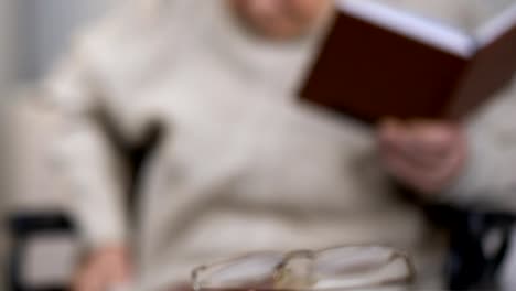 Aged-lady-in-wheelchair-holding-book-and-looking-for-eyeglasses-lying-on-table