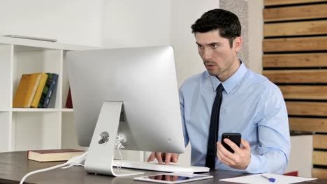 Businessman-Using-Smartphone-while-Working-on-Computer