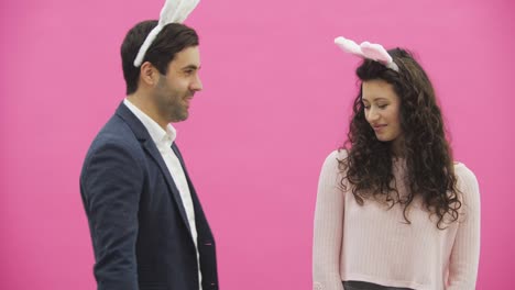 Young-creative-couple-on-pink-background.-With-hackneyed-ears-on-the-head.-During-this-man-gives-a-soft-toy-with-a-hare-to-his-wife.-Embracing-a-girl-looking-into-the-camera-and-showing-a-gesture-class.