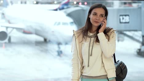 Beautiful-girl-in-sportswear-standing-at-window-in-airport-and-talking-on-phone-background-of-plane