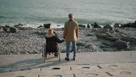 Wheelchair-woman-user-and-healthy-man-are-holding-hands-and-watching-seascape