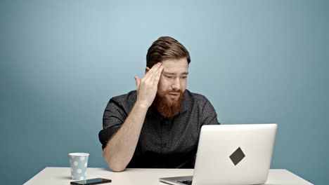 miserable-and-bored-caucasian-young-man-sitting-behind-his-office-desk-and-trying-to-work-in-front-of-a-light-blue-wall