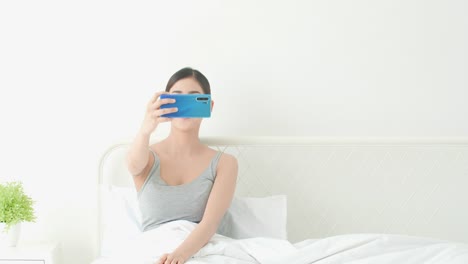 Mujer-asiática-publicando-y-tomar-fotos-para-una-nueva-foto-de-sí-mismo-con-el-teléfono-inteligente-en-la-habitación-de-la-cama.-Concepto-de-tecnología,-comunicación,-webcam,-subido-y-selfie.