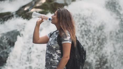 Attraktive-Mädchen-trinkt-kaltes-Wasser-aus-einer-Flasche-und-löscht-Durst,-während-auf-einem-Stein-in-der-Nähe-eines-Wasserfalls-stehen