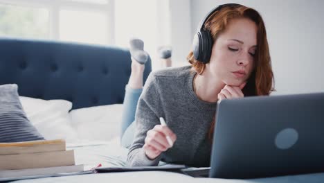 Female-college-student-lying-on-bed-wearing-headphones-and-working-on-laptop---shot-in-slow-motion