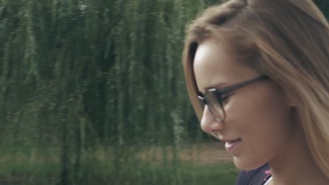 Una-mujer-caucásica-sonriente-con-gafas-camina-por-el-parque-de-la-ciudad-y-usa-su-teléfono.-Bonita-mujer-de-verano-en-blanco-t-corto-camina-por-la-calle-mirando-a-su-teléfono-móvil