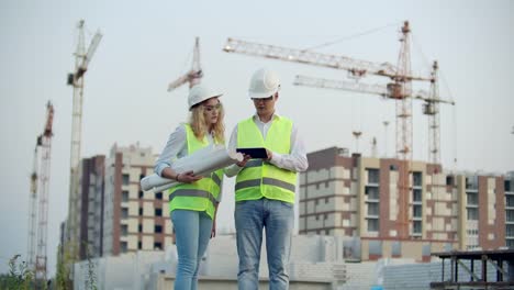 Negocios,-construcción,-industria,-tecnología-y-concepto-de-personas---constructor-sonriente-en-hardhat-con-tableta-PC-ordenador-junto-con-la-mujer-con-dibujos-de-constructores-en-el-sitio-de-construcción.