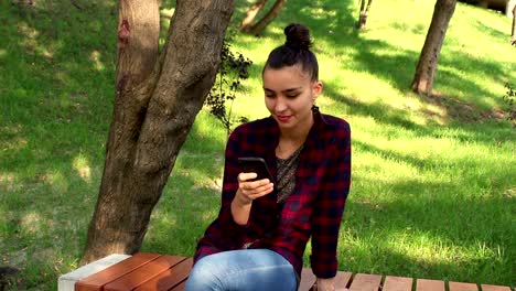 Young-beautiful-girl-in-a-plaid-shirt-sits-on-a-bench-in-the-park,-actively-rewritten-in-internet-chat