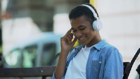 Positiver-junger-Mann,-der-Musik-in-Kopfhörern-hört-und-auf-der-Bank-sitzt