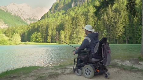 Zeitlupe-von-behinderten-Fischer-in-einem-elektrischen-Rollstuhl-Angeln-in-schönen-See-in-der-Nähe-von-Wald-und-Berg-im-Rücken