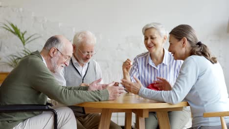 Seguimiento-de-la-toma-de-grupo-de-cuatro-jubilados-jubilados-alegres,-dos-hombres-y-dos-mujeres,-divirtiéndose-sentados-en-la-mesa-y-jugando-bingo-juntos-en-un-asilo-de-ancianos