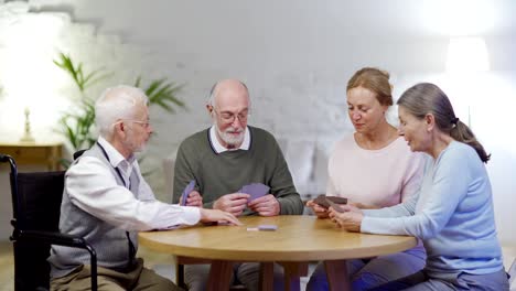 Grupo-de-cuatro-personas-mayores,-dos-hombres-incluyendo-discapacitados-uno-en-silla-de-ruedas-y-dos-mujeres,-jugando-a-las-cartas-y-hablando-sentados-a-la-mesa-en-un-asilo-de-ancianos