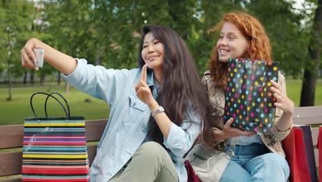 Fröhliche-Mädchen-machen-Selfie-mit-Einkaufstaschen-sitzen-auf-der-Bank-im-Park-posiert