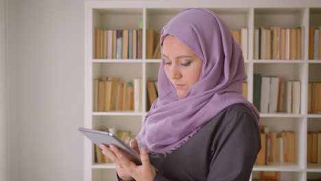 Retrato-de-primer-plano-de-joven-musulmana-en-hiyab-usando-la-tableta-y-mirando-la-cámara-de-pie-en-la-biblioteca-en-el-interior