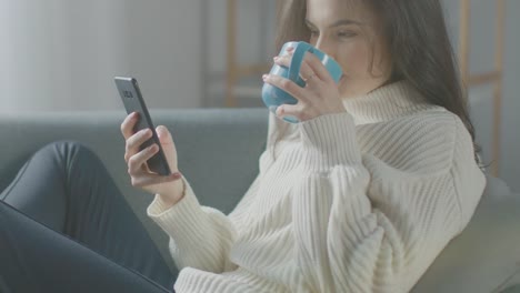 Schöne-junge-Frau-mit-Smartphone,-trinkt-Tee,-während-auf-dem-Stuhl-sitzen.-Sinnliche-Mädchen-tragen-Pullover-Surfs-Internet,-Beiträge-auf-Social-Media,-teilt-Bilder-beim-Entspannen-in-cozy-Apartment