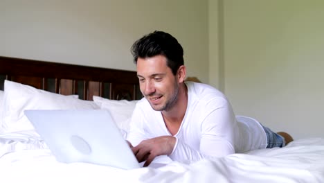 Young-Man-Using-Laptop-Computer-Lying-On-Bed-Happy-Smiling-Guy-Chatting-Online-In-Bedroom-Morning