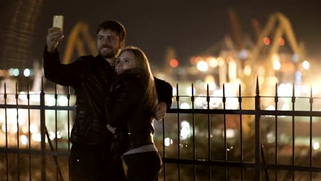 Young-happy-couple-take-selfie-photos-at-camera-on-smart-mobile-cell-phone.-Night-lights-of-port