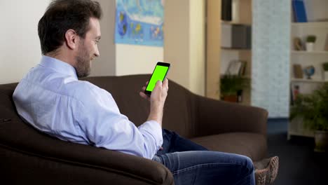 Relaxed-man-sitting-on-couch-and-viewing-photos-in-social-networks-on-cellphone