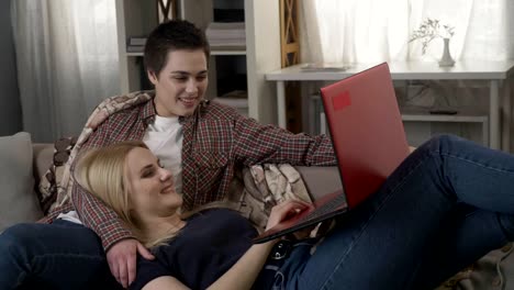 Young-blond-woman-lies-on-the-lap-of-a-brunette-and-using-red-laptop,-cosiness,-cuddles,-plaid,-talking,-dialogue-60-fps